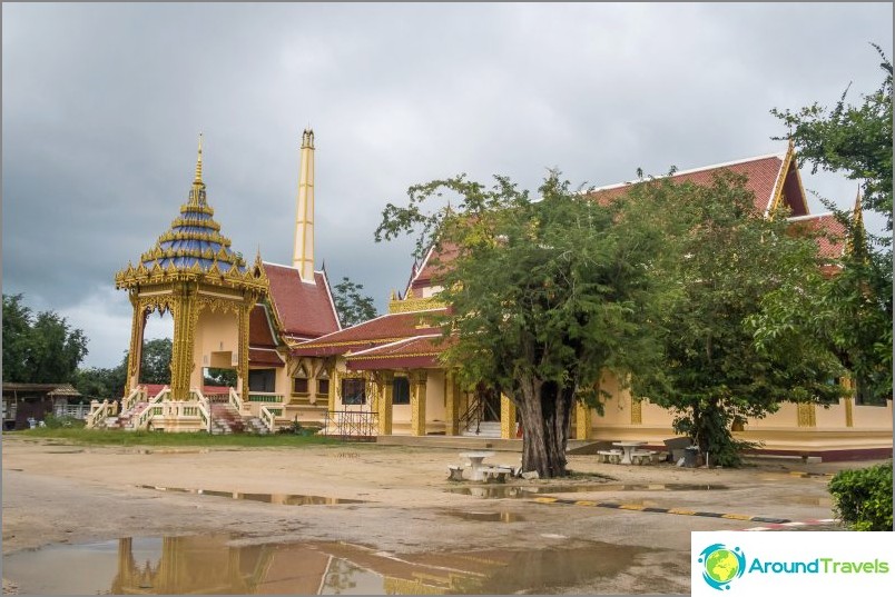 Very Thai photo: crematorium, a temple, a table under a tree, puddles and a speed bump on an earthen road.